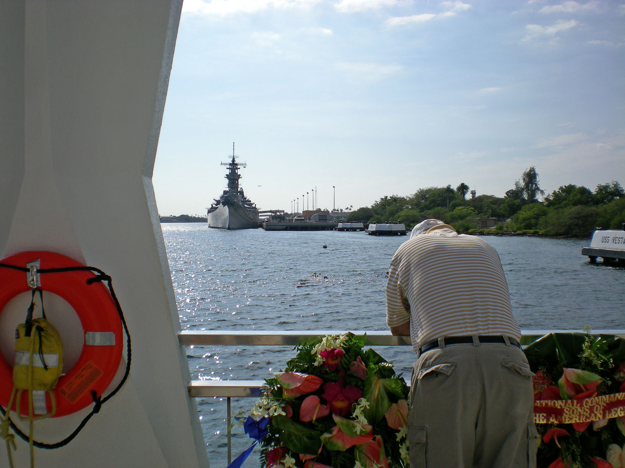 Man on boat in foreground hangs head in reverence to battleship and pearl harbor memorial in the background